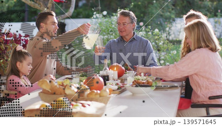 Pouring man in tan sweater lifting pitcher while family sitting on garden patio, with bread basket 139571645