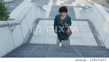 Climbing woman in teal dress ascending concrete steps to tiled plaza, holding phone and brown bag Climbing woman in teal dress ascending concrete steps to tiled plaza, holding phone and brown bag 139571837