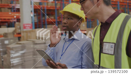 Gesturing lead in yellow hardhat holding tablet, speaking to colleague in lime vest in warehouse Gesturing lead in yellow hardhat holding tablet, speaking to colleague in lime vest in warehouse 139572100