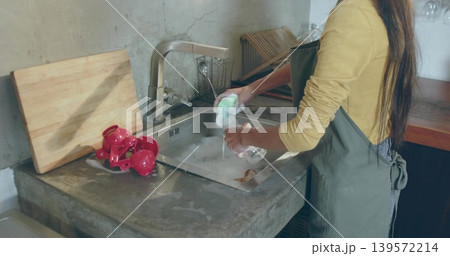 Washing woman in green apron, mustard top rinsing glass with sponge at home sink, copy space Washing woman in green apron, mustard top rinsing glass with sponge at home sink, copy space 139572214
