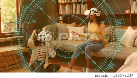 Gesturing two women wearing white VR headsets, interacting with teal holographic HUD in living room Gesturing two women wearing white VR headsets, interacting with teal holographic HUD in living room 139572234