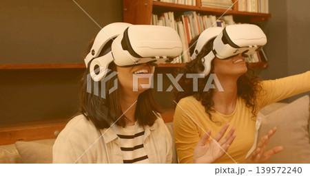 Reaching, smiling women wearing white VR headsets on sofa, striped and yellow tops near bookshelf Reaching, smiling women wearing white VR headsets on sofa, striped and yellow tops near bookshelf 139572240