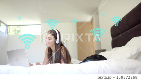 Lying woman using silver laptop on bed, wearing white top and headphones, teal Wi-Fi graphics Lying woman using silver laptop on bed, wearing white top and headphones, teal Wi-Fi graphics 139572269
