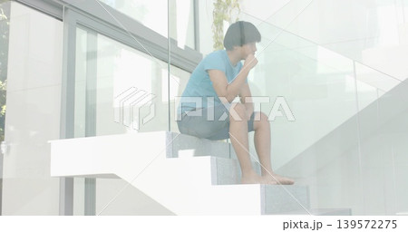 Sitting man in blue tee and denim-shorts on white floating stairs by glass with foliage reflections 139572275