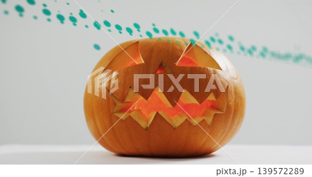 Glowing carved orange pumpkin casting warm light on white tabletop, revealing turquoise paint band 139572289