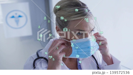 Adjusting female doctor in lab coat securing light blue mask in clinic, stethoscope, digital icons 139572536