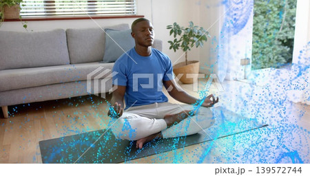Sitting man meditating in blue-tee gray-pants on yoga-mat with cushion at home with blue particles Sitting man meditating in blue-tee gray-pants on yoga-mat with cushion at home with blue particles 139572744