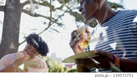 Man on right wearing navy-striped T-shirt and watch eating burger on paper plate in sunlit park Man on right wearing navy-striped T-shirt and watch eating burger on paper plate in sunlit park 139572751