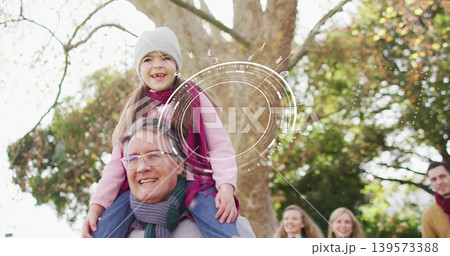 Carrying senior man holding child on shoulders at park, wearing coats and scarves, tree overlay 139573388
