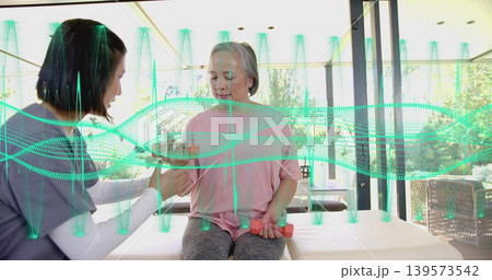 Therapist assisting woman in pink top, grey pants lifting pink dumbbells by glass walls, waveform 139573542