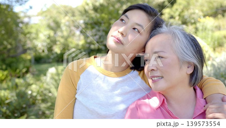 Leaning mother and daughter touching heads in sunlit park, wearing pink blouse and mustard sleeves 139573554
