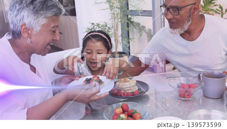 Smiling woman wearing white top offering plate with berries to child at home, pancakes on table Smiling woman wearing white top offering plate with berries to child at home, pancakes on table 139573599