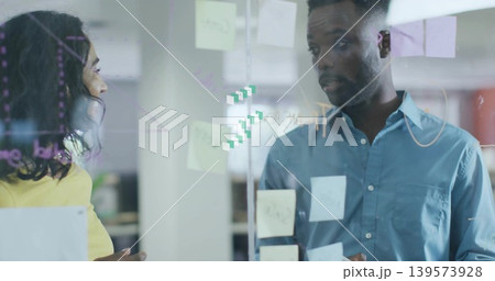 Gesturing coworkers in yellow-top and blue-shirt discussing plan in office, glass with sticky notes 139573928