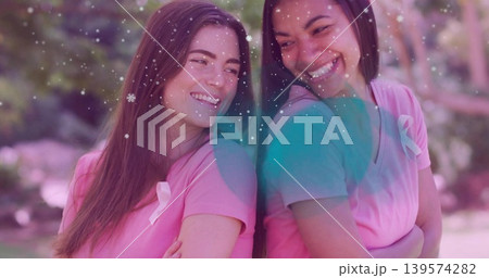 Smiling two women leaning back-to-back in park, wearing matching pink shirts with ribbons Smiling two women leaning back-to-back in park, wearing matching pink shirts with ribbons 139574282