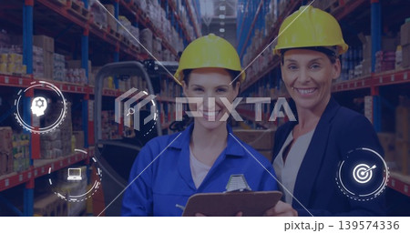 Standing two women in coveralls and blazer in yellow hard hats checking warehouse with forklift Standing two women in coveralls and blazer in yellow hard hats checking warehouse with forklift 139574336