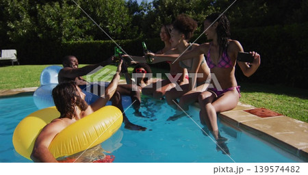 Toasting six adults clinking green bottles at backyard pool, wearing swimsuits, holding smartphone Toasting six adults clinking green bottles at backyard pool, wearing swimsuits, holding smartphone 139574482