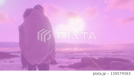 Standing couple wrapped in light blanket facing ocean on rocky shore, pink-purple sun, copy space Standing couple wrapped in light blanket facing ocean on rocky shore, pink-purple sun, copy space 139574563