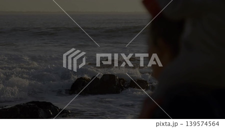 Watching woman wearing jacket, hat gazing at breaking waves on rocky shore at dusk, copy space Watching woman wearing jacket, hat gazing at breaking waves on rocky shore at dusk, copy space 139574564