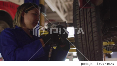 Working mechanic in blue coveralls tightening lug nuts at garage, holding impact wrench Working mechanic in blue coveralls tightening lug nuts at garage, holding impact wrench 139574623
