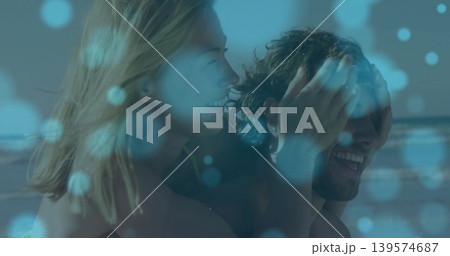 Giving woman covering man's eyes carrying him at beach shoreline, swimsuit top, trunks, blue bokeh Giving woman covering man's eyes carrying him at beach shoreline, swimsuit top, trunks, blue bokeh 139574687
