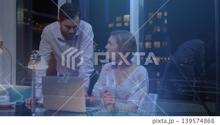 Discussing team in business attire leaning over glass table with laptop overlay in office at night 139574868