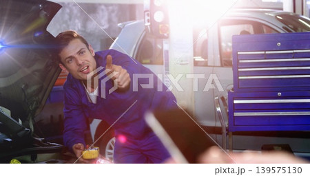 Leaning mechanic in blue coveralls checking open hood in garage, holding yellow tool near phone 139575130