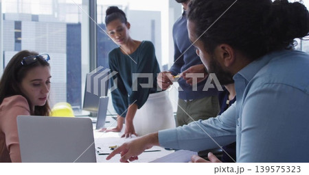 Pointing man with bun wearing blue shirt leaning over table in modern office, laptop and sketches Pointing man with bun wearing blue shirt leaning over table in modern office, laptop and sketches 139575323