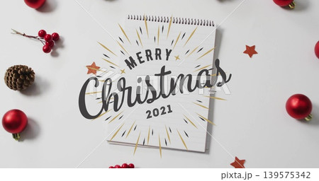 Displaying spiral notepad featuring Merry Christmas 2021 on white tabletop, with baubles, pinecone 139575342