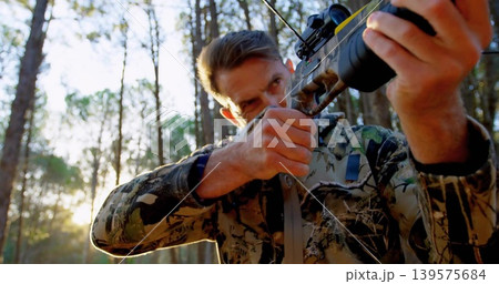 Aiming mature hunter steadying crossbow with mounted scope in woods, wearing camouflage jacket 139575684