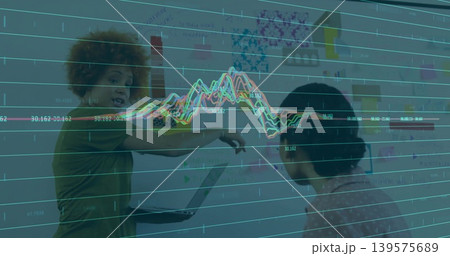 Pointing two women working at office wall with open laptop sticky notes, green top, printed blouse 139575689