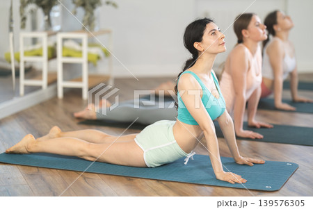 Women bending in back while lying on stomach in Bhujangasana Cobra Stretch position during training together in studio 139576305
