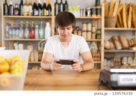 Bored guy seller with mobile phone in hands plays online games, spends time online Bored guy seller with mobile phone in hands plays online games, spends time online 139576339