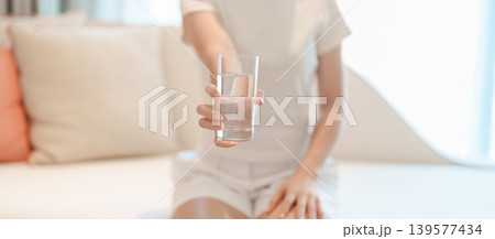 Healthy woman hold glass of clean mineral water glass on sofa at home, female drink pure water for hydration and wellness, fresh skin care, daily routine water intake, Refreshment and lifestyle 139577434