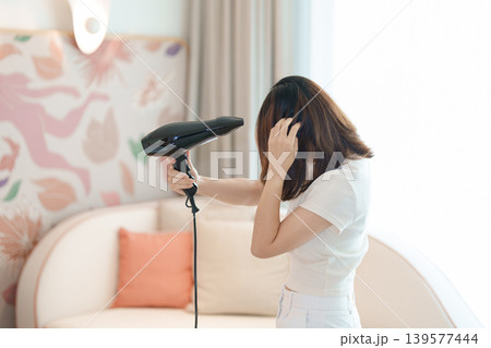 Young woman drying long hair with a hair dryer after a shower in home or hotel room during vacation. Hairstyles and daily morning routine Young woman drying long hair with a hair dryer after a shower in home or hotel room during vacation. Hairstyles and daily morning routine 139577444