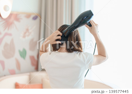 Young woman drying long hair with a hair dryer after a shower in home or hotel room during vacation. Hairstyles and daily morning routine 139577445