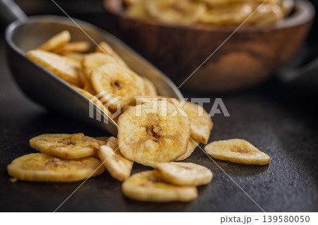 Crispy Banana Chips in Metal Scoop and Wooden Bowl on Dark Surface. Dried Fruit Snack Crispy Banana Chips in Metal Scoop and Wooden Bowl on Dark Surface. Dried Fruit Snack 139580050