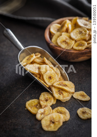Crispy Banana Chips in Metal Scoop and Wooden Bowl on Dark Surface. Dried Fruit Snack 139580051