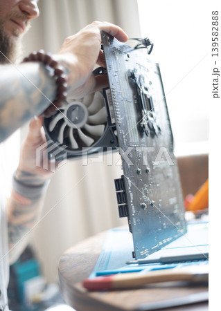 Person repairs computer hardware with a cooling fan and circuit board at a table during daytime in a minimalistic room setting Person repairs computer hardware with a cooling fan and circuit board at a table during daytime in a minimalistic room setting 139582888