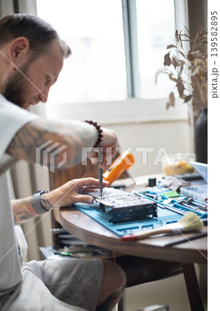 Man works on repairing electronics at home during the day with tools and materials scattered around the table 139582895