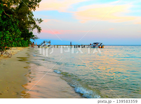 Sunset view of wooden pier with motor boats and people enjoying beach at an ocean shore Sunset view of wooden pier with motor boats and people enjoying beach at an ocean shore 139583359