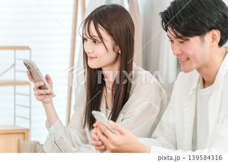 家のリビングでソファーに座りスマホを見ながら会話する若いアジア人夫婦 139584316