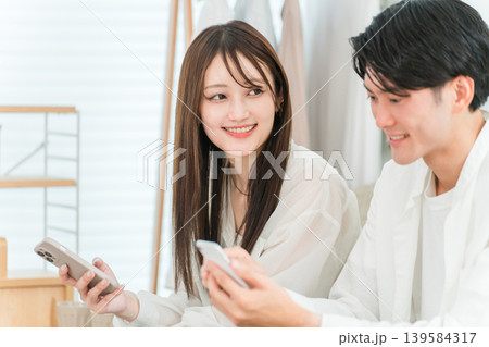 家のリビングでソファーに座りスマホを見ながら会話する若いアジア人夫婦 139584317
