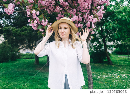 Beautiful young woman in straw hat posing near blooming pink sakura tree in spring park. Romantic girl enjoying cherry blossom outdoors. 139585068