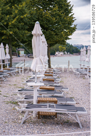 Rows of empty sunbeds and closed umbrellas on pebble beach, peaceful lakeside atmosphere with trees and soft cloudy sky in background 139586729