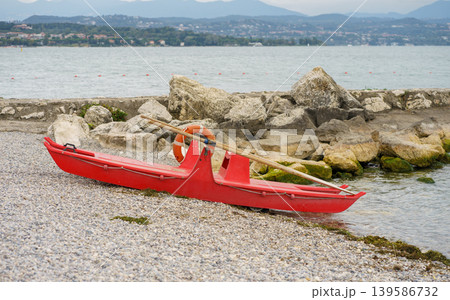 Red catamaran boat with oars and lifebuoy on pebble shore, calm water and rocky coastline create peaceful lakeside scene with natural textures Red catamaran boat with oars and lifebuoy on pebble shore, calm water and rocky coastline create peaceful lakeside scene with natural textures 139586732