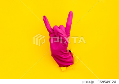 Pink cleaning glove rock gesture through torn yellow paper background Pink cleaning glove rock gesture through torn yellow paper background 139589128