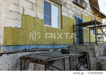 Workers are applying insulation to a house wall while using a scaffold system. Tools and materials are placed nearby on a wooden table. Wall insulation. 139589995