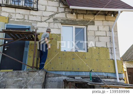 A man is applying insulation panels to the wall of a house while standing on a scaffold. He works to improve the home's energy performance. Wall insulation A man is applying insulation panels to the wall of a house while standing on a scaffold. He works to improve the home's energy performance. Wall insulation 139589996
