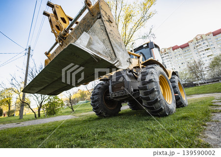 A construction vehicle is lifting dirt in a park setting. Trees are visible around, and buildings are seen in the background under clear skies. Loader A construction vehicle is lifting dirt in a park setting. Trees are visible around, and buildings are seen in the background under clear skies. Loader 139590002