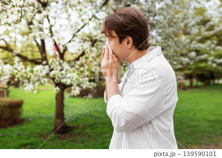 A man sneezes into a tissue while standing near blooming trees in a park. This image depicts seasonal allergies, hay fever, and the impact of spring pollen on health. A man sneezes into a tissue while standing near blooming trees in a park. This image depicts seasonal allergies, hay fever, and the impact of spring pollen on health. 139590101